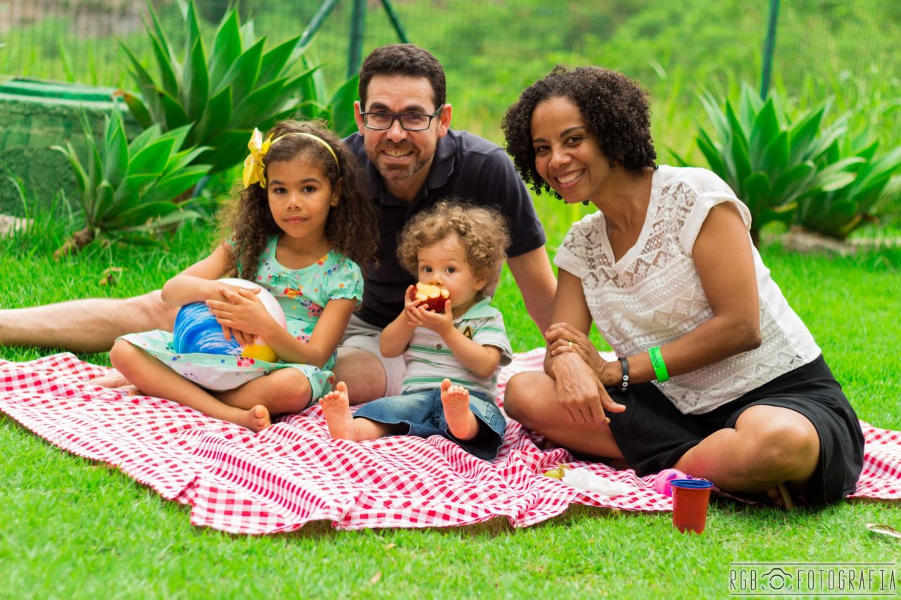Família sentada na grama em toalha de piquenique: Pai, mãe, manina e menino.