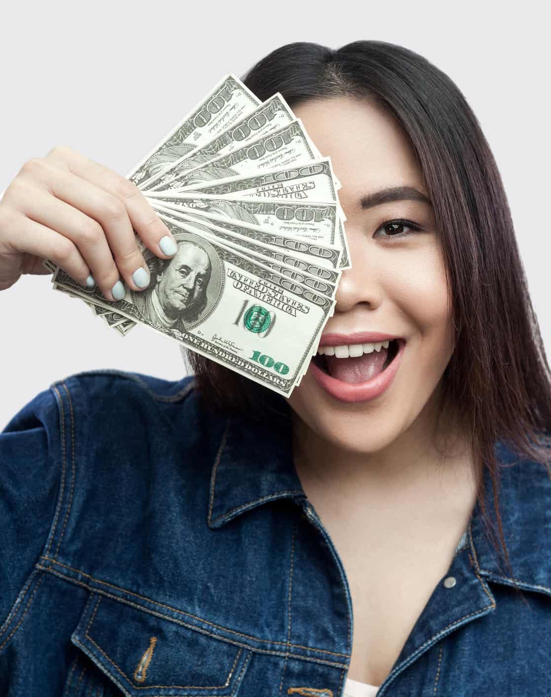 Portrait of beautiful happy young asian girl in blue denim jacket standing, holding many cash dollars and covering half of face with toothy smile. Indoor, isolated, studio shot, grey background