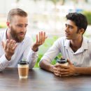 Two male friends drinking coffee and talking in outdoor cafe. People sitting at table with blurred view in background. Coffee break concept. Front view.