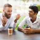 Two male friends drinking coffee and talking in outdoor cafe Two male friends drinking coffee and talking in outdoor cafe. People sitting at table with blurred view in background. Coffee break concept. Front view.